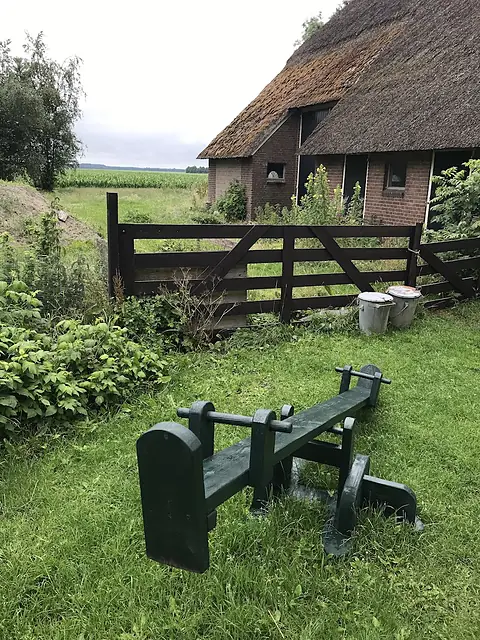Het erf met een houten wip, het hek en de boerderij met rieten dak